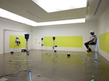 Two people sit on umpire chairs on either side of a brightly lit gallery. Sprawled upon the gallery floor are tennis ball shooter machines, tennis balls are dotted throughout then space.