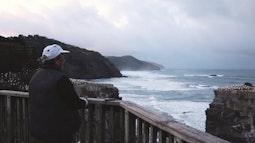 A person stands by a fence at the edge of a cliff looking out towards a rough coastline.