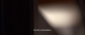 An indistinct blur of white light next to a block of black. At the bottom of the screen is a caption which reads "I am but a conversation..."