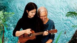 A person sits on their parents knee while playing the ukelele. A bright blue body of water is super-imposed as the background.