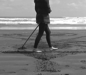 Sand is being drawn on by the artist holding a rake and dragging it across the beach. The artists head is cut off from the image but we can see their body and bare feet.