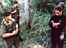 Two groups of men stand on either side of barbed wire in a forest. One side wears military uniforms.