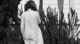 A person stands facing a rosemary bush by a house in a white dress.