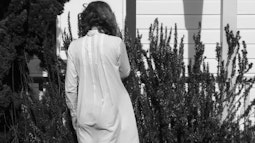 A person stands facing a rosemary bush by a house in a white dress.