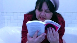 A person in a red coat sits in a bath reading a book.