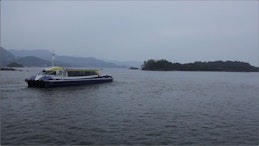 A short and long ferry floats on a body of water on a dark and cloudy day. The ferry sits on water next to a small island in the background.