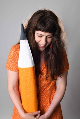 A woman with brown hair wearing an orange dress and holding a large fake pencil.