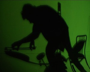 The silhouette of a figure standing against a green background. A chair is also silhouetted and other instruments.