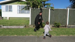 A person runs and plays with a small child on a pavement. There is a house and tall fence behind them.