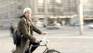 A person on a bike in a city looks into the camera, the background is motion blurred and the figure is in focus.