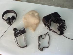Headphones, oculus, camera, goggles and a plastic face are laid out on a clean white table