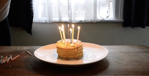 A stack of small pancakes decorated with birthday candles is dripping with honey on na plate. The lit pancakes sit alone on a table in a flat, behind the table is a window with netted curtains.