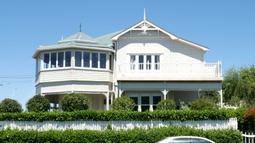 A large white house stands tall above a green hedge row.