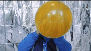 A giant yellow balloon is being blown up by a person wearing bright blue standing infront of metalic silver backrgound