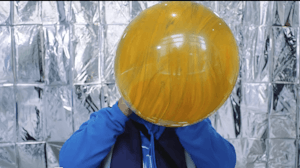 A giant yellow balloon is being blown up by a person wearing bright blue standing infront of metalic silver backrgound
