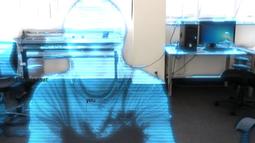 A person appears like a hologram in an office, they're made of translucent blue light.