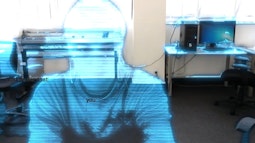 A person appears like a hologram in an office, they're made of translucent blue light.