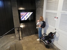 A person stands next to a stroller, holding a small baby and watching the video work playing on masons screen