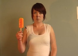 Erica holds up a bright orange popsicle in front of a plain wall. They wear a white singlet and a deadpan facial expression