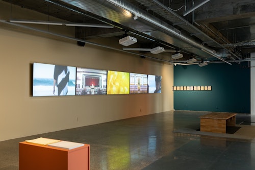 A gallery with a wall of five large LCD screens, showing images of people by the ocean, and individuals addressing rooms suggesting highly formal or political settings. At right a line of government documents are framed as individual sheets of A4 paper. Some are torn.