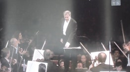 A conductor stands in front of an orchestra wearing a suit