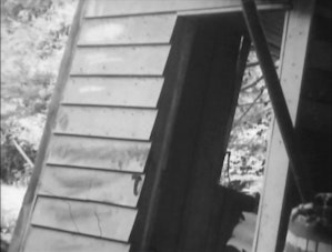 A grainy black and white image of a small house of shed with the door open. The house is made from weatherboards and inside looks like things have fallen over.