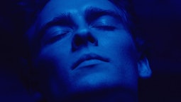 A close-up of a young man with his eyes closed in pleasure, he is cast in a deep blue light.
