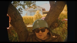 A man leans against a tree wearing a army green helmet and sunglasses.