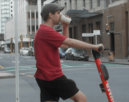 Max is on a segway, waiting to cross the street, drinking out of a white takeaway cup.