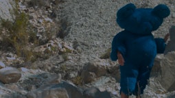 A person wears a large blue furry teddy bear costume as they walk amongst dry rocks.