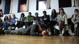A person lies on the ground with a toilet plunger as a row of people look on.