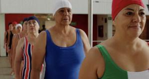 A line of older women in swimsuits and swim-caps walk along the perimiter of the pool, some of them look excited and some look a little nervous