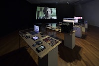 Museum-esque display of objects and monitors inside glass vitrines