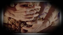 A close up of two hands clutching a string of beads and shells. Linear tattoos are visible on the figure's fingers and wrists