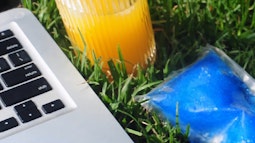 A close up of glass of orange juice, an ice pack, and a laptop on a patch of grass.