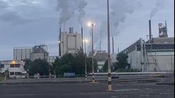 A large factory with smoke stacks billows gasses into the air. Flood lights are in the foreground shining over bare concrete.