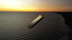A digitally rendered metal oblong flies above the sea while the sun sets.