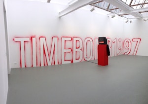The text TIMEBOMB1997 is spraypainted on the wall of the gallery, in front of this is a red plinth with a black monitor on top
