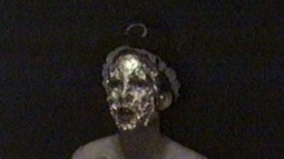 A person who's face is covered in gold looks just out of frame. They are wearing a crown and stood against a black backdrop.