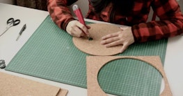 A person sits at a desk and uses a craft knife to carve a circle out of a cork board.