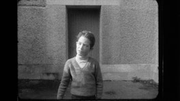 A young boy stands in front of a door way talking, he is dressed in a knitted sweater and turtleneck.