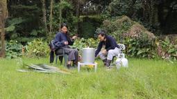 Two people sit in plastic chairs on a lush patch of grass. Dense bush surrounds them. There is a large metal pot and gas canister between them.