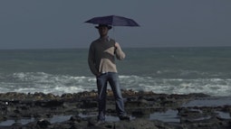 A person stands on rocks at the coast holding an umbrella above their head.