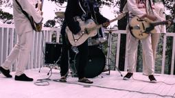 A four person band plays on a white wood deck. Three people are playing guitars and one is playing the drums.