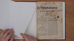 A birds-eye view of someone's hands turning pages in a book. The heading of the book reads "Te Pipiwharauroa"