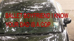 A car parked beneath a tree with dappled light reflections. Large text on screen reads, Ballet boyfriend I know your dad is a cop.