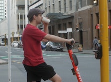 Max sips out of a cup whilst waiting at a pedestrian crossing in Wellington Central. He is holding an electric scooter in anticipation for the light to change colours and to ride off. Max wears a brown hat, red tshirt and black shorts.