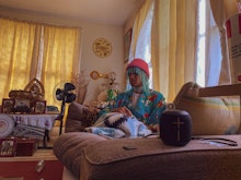 A person sits in a lounge room on a comfortable couch wearing a pink beanie with jewels hanging out of from the beanie. They are listening to something on their Bluetooth speaker