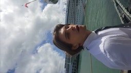 A person looks at the camera with their mouth slightly agape, they're wearing a white shirt and tie. There is a large cruise ship in behind them.
