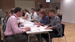Participants in a performance sit at a long table in a conference room folding paper